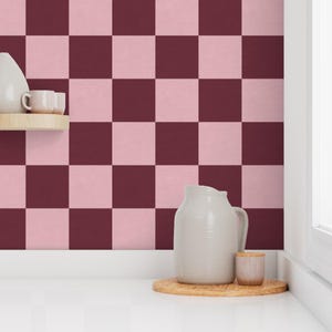 Puede incluir: Un patr&oacute;n de tablero de ajedrez en tonos rosa y burdeos cubre la pared. Una jarra blanca, una peque&ntilde;a taza de madera y una bandeja de madera se encuentran sobre una superficie blanca. Una peque&ntilde;a estanter&iacute;a contiene un jarr&oacute;n blanco y dos tazas peque&ntilde;as.