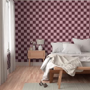 Puede incluir: Un dormitorio con una pared a cuadros burdeos y rosa. La habitaci&oacute;n tiene una mesita de noche de madera con una l&aacute;mpara, una cama con ropa de cama blanca y un banco de madera al pie de la cama. El suelo es de madera clara y se ve una ventana con cortinas blancas.