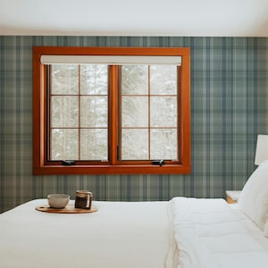 May include: A bedroom interior featuring walls covered in a blue and grey plaid wallpaper. A wooden window frame surrounds a double-pane window. A white bed with a white comforter and pillows sits in the foreground with a small wooden tray.