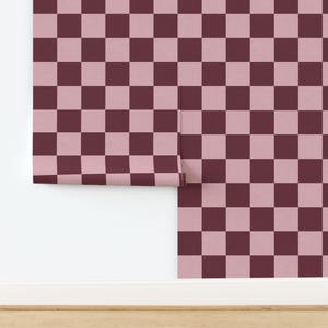Puede incluir: Rollo de papel pintado con un patr&oacute;n de tablero de ajedrez repetido en tonos burdeos y rosa claro. El papel pintado est&aacute; parcialmente desenrollado, revelando el patr&oacute;n contra una pared blanca y un suelo de madera.