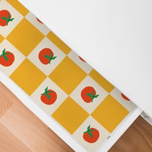 May include: A drawer interior lined with a patterned paper featuring a repeating design of red tomatoes with green stems on a yellow and cream checkered background. The drawer is white, and the floor is wood.