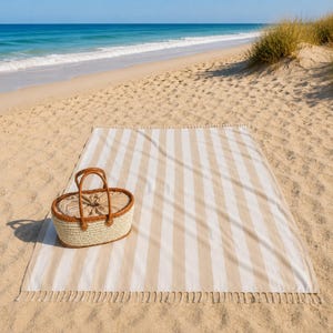 Op de afbeelding: Een beige en wit gestreepte stranddeken met franjes en een rieten picknickmand met een leren handvat liggen op een zandstrand. De oceaan en de blauwe lucht zijn op de achtergrond te zien. Ideaal voor een dagje uit.