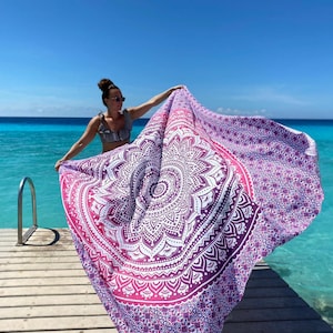 May include: A large, round beach towel with a mandala design. The towel features a gradient of colours, transitioning from white to pink and purple. The intricate pattern is centered on the towel, with a repeating design around the edges. The towel is being held up near a body of water.