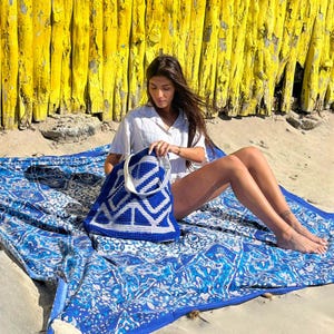 Op de afbeelding: Een vrouw zit op een blauw-witte gebloemde deken met een blauwe en witte gebreide draagtas. De deken ligt op een zandstrand voor een gele houten schutting.