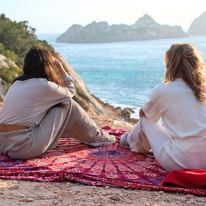 May include: A red and purple mandala patterned beach blanket is spread on a rocky cliff overlooking the ocean. Two people are seated on the blanket, facing the water. A red hat is placed on the blanket.