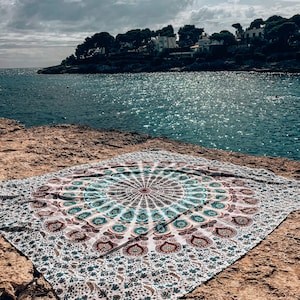 Puede incluir: Un tapiz blanco con un diseño de mandala colorido que presenta tonos de azul, verde y rosa. El tapiz está colocado sobre una superficie rocosa cerca del océano.