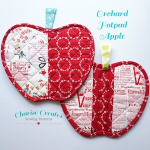 May include: A pair of red and white quilted apple-shaped hot pads with a floral print. The hot pads are attached together with a green and white fabric loop. The text "Orchard Hotpad Apple" is printed above the hot pads.