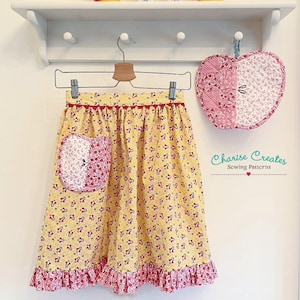Puede incluir: Un delantal de estampado floral amarillo y rojo con un dobladillo con volantes y un bolsillo rojo en forma de manzana. El delantal está colgado de una percha de madera. Hay un posavasos rojo en forma de manzana a juego colgado al lado del delantal. El texto "Charlise Creates Sewing Patterns" es visible en la esquina inferior derecha de la imagen.