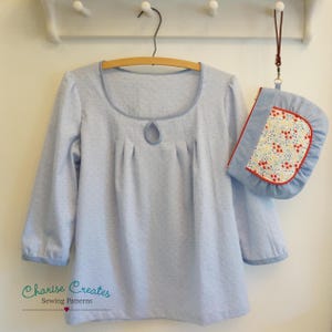 May include: A light blue, long-sleeved blouse with a keyhole neckline and a gathered bodice. The blouse is hanging on a wooden hanger. A small, floral-patterned pouch with a red trim is hanging next to the blouse.