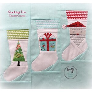 May include: A Christmas stocking trio quilt block with a green, white, and red stocking, a blue and white stocking with a present, and a gray stocking with a red hat and the words "present box". The background is a light blue color.