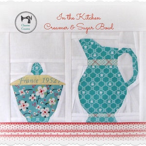 May include: A white fabric background with a blue and white patterned creamer and sugar bowl set. The creamer has a handle and the sugar bowl has a lid with the text "France 1952". The set is on a red and white striped fabric.