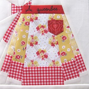 May include: A red, white, and yellow patchwork quilt block with a red gingham border. The block features a floral print and a red pocket with a heart design. The text "A queenbee." is stitched in red on the top of the block.