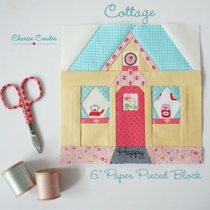 Puede incluir: Un bloque de acolchado de papel de 15 cm que presenta una cabaña amarilla con una puerta y ventanas rosas. La cabaña tiene un techo a cuadros azul y blanco y un borde floral rosa y blanco. El texto "Cottage" está escrito en turquesa sobre el bloque y "Happy" está escrito en negro debajo del bloque.