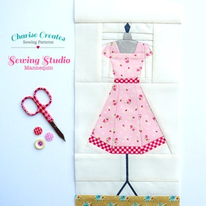 May include: A pink dress with a floral print and a red and white gingham trim is displayed on a white mannequin. The dress is part of a sewing studio themed quilt block.