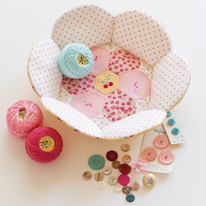 May include: A flower-shaped fabric basket with a pink polka dot exterior and a patchwork interior featuring strawberries and cherries. Pink and teal thread spools and various buttons are scattered around the basket, perfect for sewing and crafting.