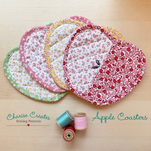 May include: Four heart-shaped quilted pot holders in green, yellow, pink, and red floral fabric with a yellow, pink, or green trim. The pot holders are arranged in a stack on a wooden surface. Three spools of thread are in the lower right corner of the image. The text "Apple Coasters" is in the lower right corner of the image.