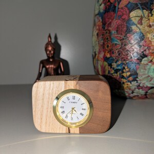 Op de afbeelding: Een houten klok met een gouden wijzerplaat en Romeinse cijfers. De klok staat op een houten voet met licht hout aan de ene kant en donker hout aan de andere kant. De wijzerplaat van de klok geeft "CAMRY QUARTZ" aan.
