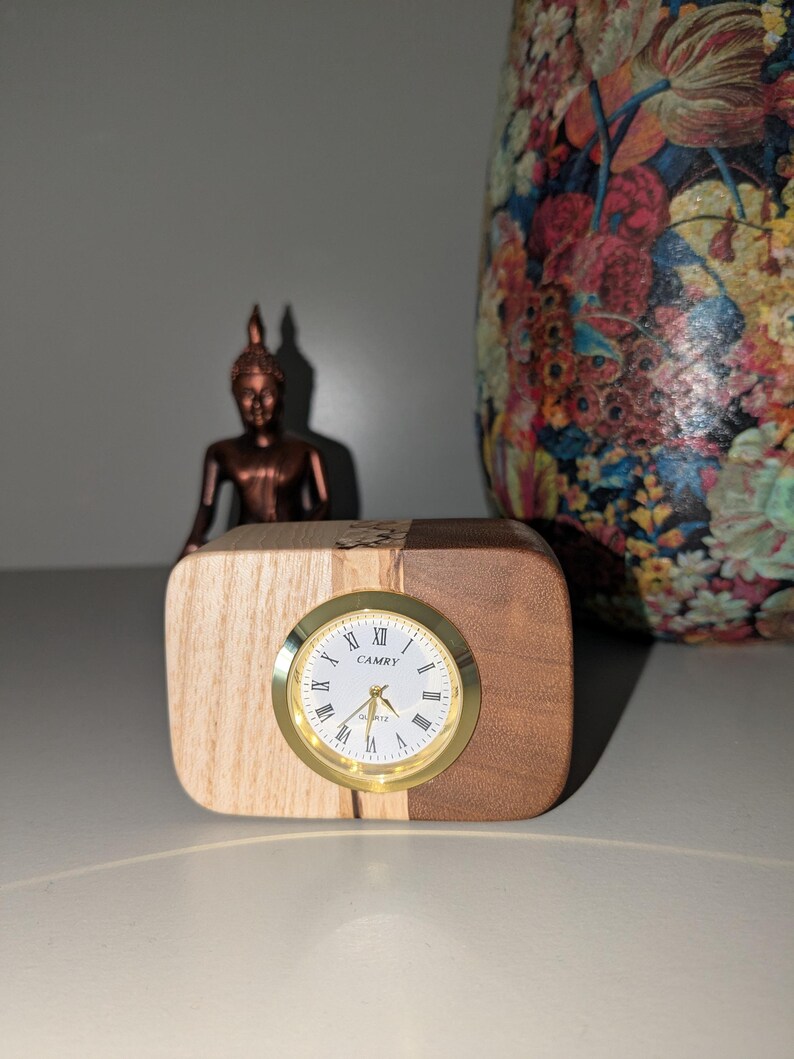 Op de afbeelding: Een houten klok met een gouden wijzerplaat en Romeinse cijfers. De klok staat op een houten voet met licht hout aan de ene kant en donker hout aan de andere kant. De wijzerplaat van de klok geeft "CAMRY QUARTZ" aan.