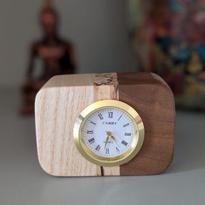 Op de afbeelding: Een houten klok met een gouden wijzerplaat en Romeinse cijfers. De wijzerplaat van de klok geeft "CAMRY QUARTZ" aan.