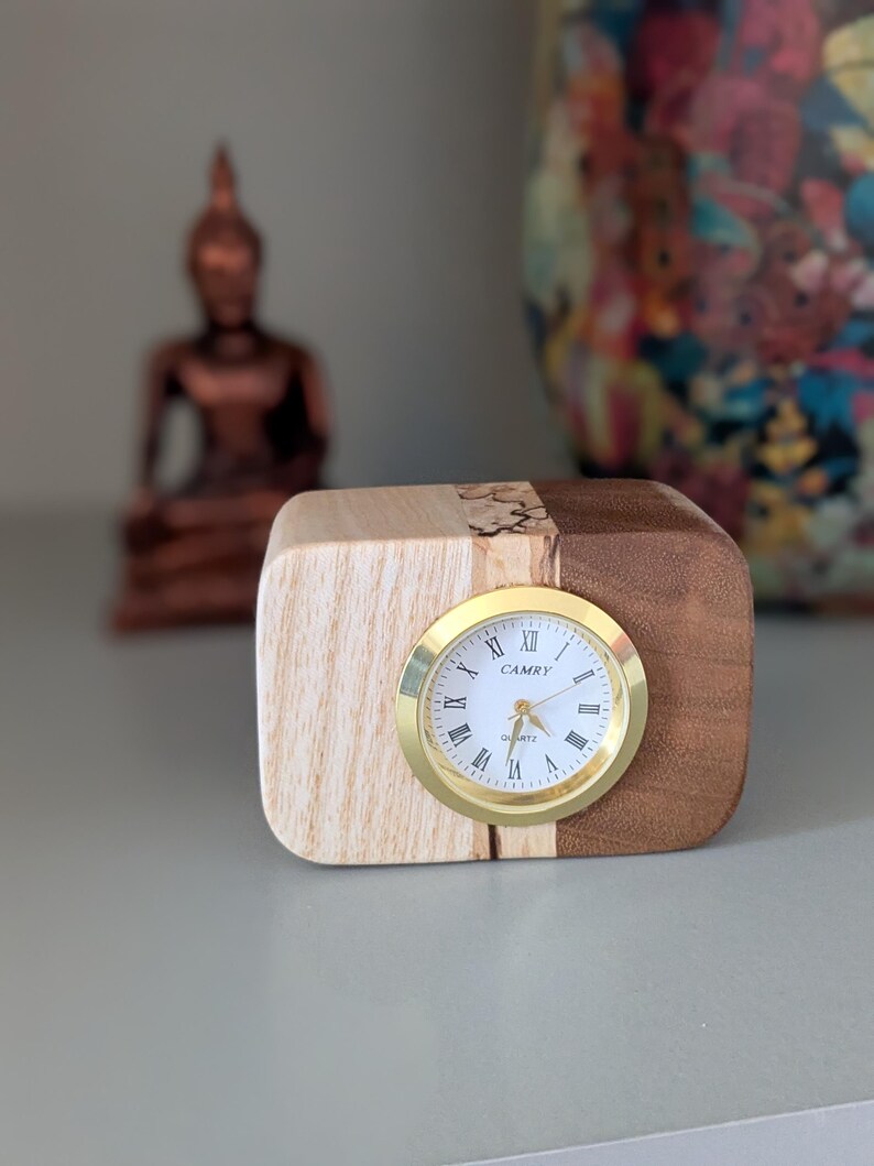 Op de afbeelding: Een houten klok met een gouden wijzerplaat en Romeinse cijfers. De wijzerplaat van de klok geeft "CAMRY QUARTZ" aan. De klok staat op een houten voet met licht hout aan de ene kant en donker hout aan de andere kant.
