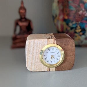 Op de afbeelding: Een houten klok met een gouden wijzerplaat en Romeinse cijfers. De wijzerplaat van de klok geeft "CAMRY QUARTZ" aan. De klok staat op een houten voet met licht hout aan de ene kant en donker hout aan de andere kant.