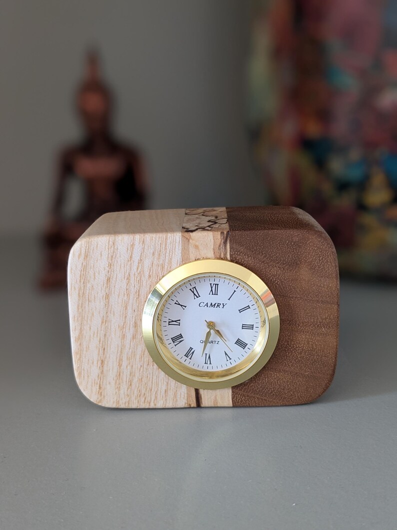 Op de afbeelding: Een houten klok met een gouden wijzerplaat en Romeinse cijfers. De wijzerplaat van de klok geeft "CAMRY QUARTZ" aan.