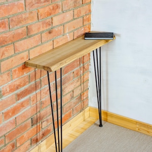 Può includere: Un tavolo consolle stretto con un piano rettangolare in legno marrone chiaro con venature naturali e quattro sottili gambe a forcina in metallo nero. Un sottile taccuino rettangolare nero è appoggiato sulla superficie del tavolo.