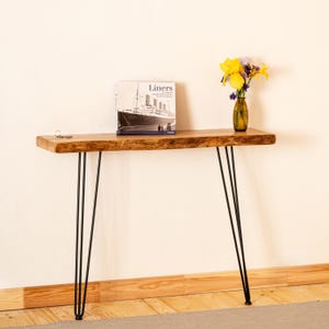 Può includere: Una consolle rustica con gambe a forcina nere. Il piano in legno naturale sostiene un libro intitolato "Liners" e un piccolo vaso con fiori gialli e viola. Una chiave è appesa al bordo. La lunghezza del tavolo è di circa 120 cm.