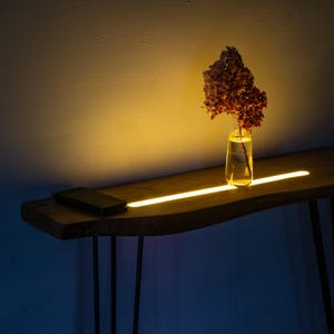 Op de afbeelding: Een houten console met een live edge design, met een ingebouwde lineaire verlichting. Een heldere glazen vaas met gedroogde bloemen staat op de tafel. Een zwarte smartphone ligt aan de linkerkant van de tafel. De tafel heeft zwarte metalen haarspeldpoten.