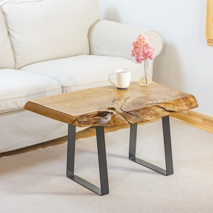 Puede incluir: Mesa de centro de madera natural con borde vivo. La mesa tiene un acabado marrón claro y descansa sobre patas de metal gris oscuro. Una taza blanca y un jarrón con flores rosas están sobre la mesa.