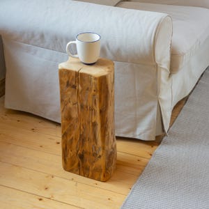 Puede incluir: Una rústica mesa auxiliar de madera, hecha de un tronco de árbol, con una taza blanca con borde azul. La mesa está junto a un sofá blanco. Dimensiones aproximadas: 30 cm x 30 cm x 50 cm.