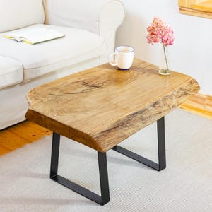 May include: A rectangular side table with a natural live edge light brown wood top featuring visible grain and a knot, supported by two black metal U-shaped legs. A white ceramic mug and a small glass vase with pink flowers sit on the table.