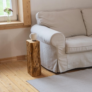 Puede incluir: Una mesa auxiliar cuadrada de madera rústica con un acabado natural y texturizado. Una taza blanca con borde negro está encima. La mesa está junto a un sofá de color claro. La mesa mide unos 40 cm de alto.