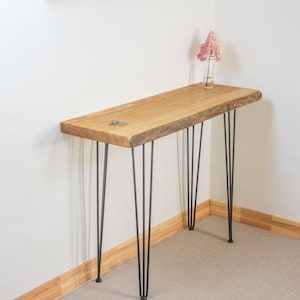 May include: A natural wood console table with a live edge top. The table has black hairpin legs and is approximately 48 inches long. A small vase with pink flowers and a set of keys rest on the table.