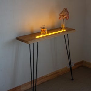 May include: A wooden console table with black hairpin legs. The tabletop has a built-in yellow light strip and holds a perfume bottle and a vase with dried flowers. The table is against a white wall.