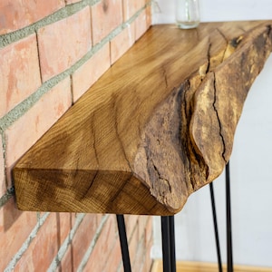 Op de afbeelding: Een console-tafel van hout met een natuurlijke boomstamrand, voorzien van een dik eikenhouten blad met een natuurlijke nerf en een ruwe, getextureerde schorsrand. De tafel wordt ondersteund door dunne, zwarte metalen haarspeldpoten.