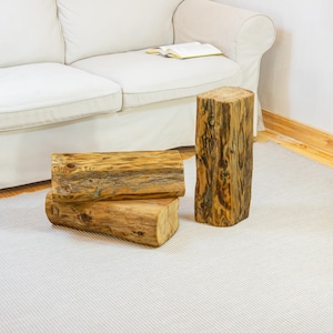 Puede incluir: Tres taburetes de tronco de madera de diferentes tamaños. Dos están apilados horizontalmente y uno está en vertical. Los troncos tienen un color marrón claro natural con vetas y nudos visibles. Se colocan sobre una alfombra gris claro cerca de un sofá blanco.