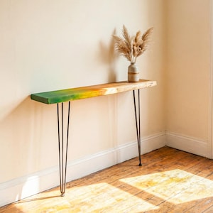 Pode incluir: Uma mesa consola estreita com um tampo de madeira de borda natural apresentando um gradiente de tons verdes, amarelos e madeira natural. A mesa é suportada por quatro pernas finas de metal preto em forma de gancho. Um pequeno vaso de cerâmica com erva-das-pampas seca está colocado em cima.