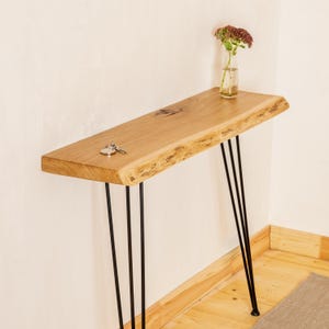 May include: A natural wood console table with a live edge and black hairpin legs. The table has a light brown finish and is approximately 48 inches long. A small vase with flowers and a set of keys rest on the table.