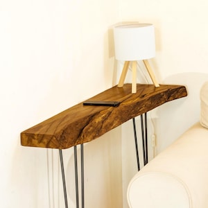 Op de afbeelding: Een console-tafel van hout met een natuurlijke rand, een donkerbruine afwerking en zichtbare houtnerf, ondersteund door vier dunne zwarte metalen haarspeldpoten. Een kleine witte lamp met een houten statief en een zwarte smartphone staan erop.