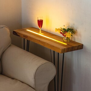 Könnte beinhalten: Ein Beistelltisch aus Holz mit einer rohen Kante und einem integrierten Leuchtstreifen. Ein Glas mit roter Flüssigkeit steht auf dem Tisch, neben einer kleinen Vase mit gelben und weißen Blumen. Der Tisch hat schwarze Haarnadelbeine aus Metall.