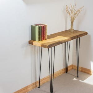 Könnte beinhalten: Eine Holzkonsolentisch mit natürlicher Kante und schwarzen Haarnadelbeinen. Auf dem Tisch stehen ein Stapel Bücher und eine kleine Glasvase mit getrocknetem Weizen. Der Tisch ist etwa 122 cm lang.