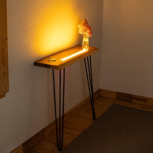 Puede incluir: Una consola de madera con una tira de luz cálida y brillante incrustada en la encimera. La mesa tiene patas de horquilla negras y un pequeño jarrón con flores secas. Un juego de llaves descansa sobre la superficie de la mesa.