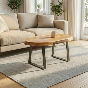 Peut inclure: Une table basse ovale avec un plateau en bois clair naturel au grain apparent, doté d'un bord brut et d'une surface lisse. La table repose sur deux pieds en métal gris foncé en forme de U. Une tasse en céramique blanche est posée au centre.