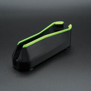 May include: A black plastic tool with a bright green trim. The tool has a U-shaped design with a rectangular body and is angled slightly upward. The green trim outlines the top edges of the tool.