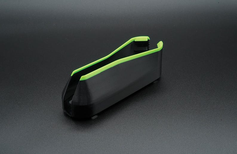 May include: A black plastic tool with a bright green trim. The tool has a U-shaped design with a rectangular body and is angled slightly upward. The green trim outlines the top edges of the tool.