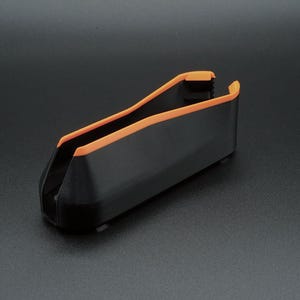 May include: A black plastic object with orange trim. The object has a curved, open design, and appears to be a holder or container. The orange trim outlines the top edges. The object is on a dark surface.
