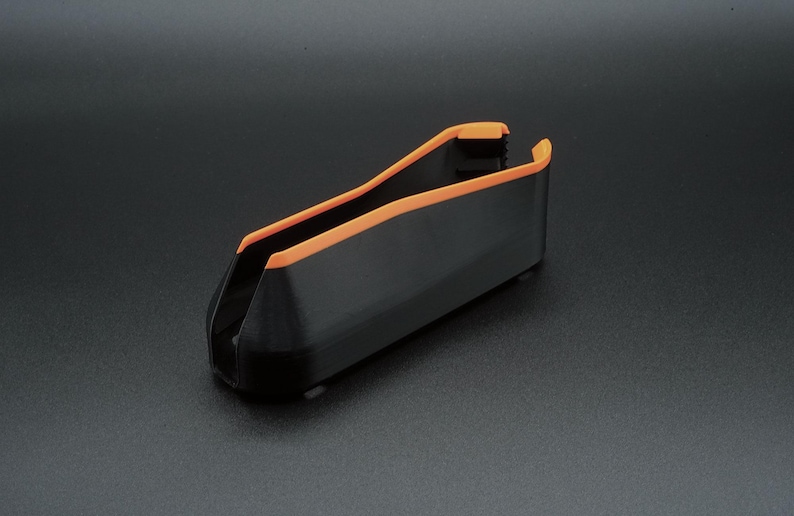 May include: A black plastic object with orange trim. The object has a curved, open design, and appears to be a holder or container. The orange trim outlines the top edges. The object is on a dark surface.