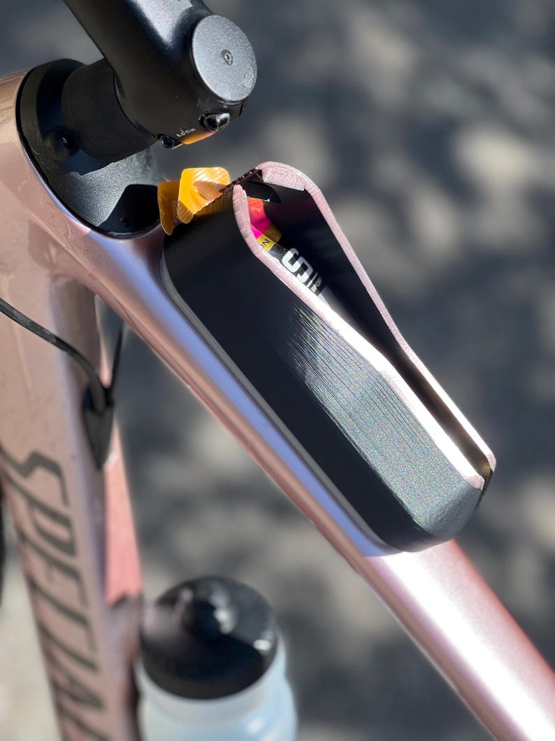 May include: A rose gold bicycle frame with a black storage compartment integrated into the frame. The compartment is open, revealing a partially visible energy gel packet and a nutrition bar. The Specialized logo is visible.