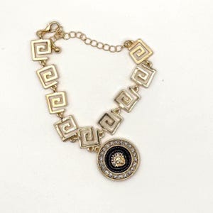 Puede incluir: Pulsera de oro con cadena cuadrada y un colgante redondo con un diseño negro y dorado. El colgante está adornado con brillantes transparentes.